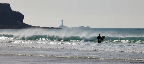 This landscape photograph captures a surfer holding a surfboard, standing in the surf at Sennen Cove beach on the coast of Cornwall in the United Kingdom. Taken in the afternoon during the spring season, the image shows natural waves rolling toward the shore with a rugged rocky coastline on the left. In the distance, the Longships Lighthouse is clearly visible offshore, acting as a notable maritime landmark. The setting emphasizes the natural beauty of the area, with the bright spring sunlight illuminating the water and the beach, characteristic of the coastal environment around Sennen Cove in Cornwall.
